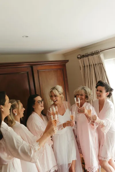 Bridal party getting ready shares a getting ready toast, smiling as they clink champagne flutes by a window before the ceremony.