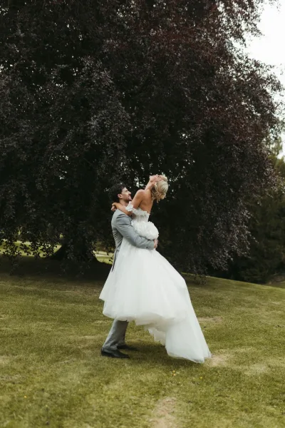 Couple portrait with groom lifting bride beneath a large tree, her bridal gown flaring as they pose on a grass lawn framed by lush greenery.