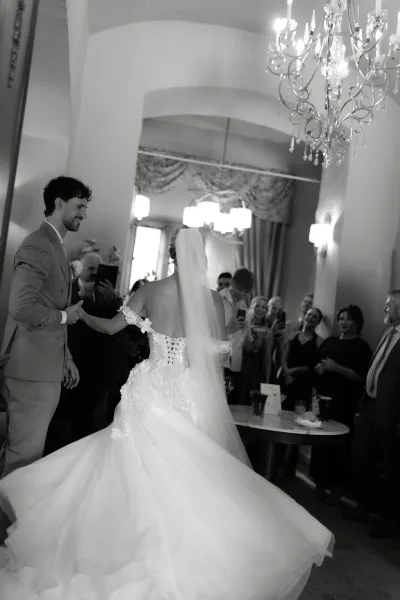 Wedding first dance with bride twirling dress as the groom holds her hand under a chandelier, framed by an arched ceiling and draped curtains.