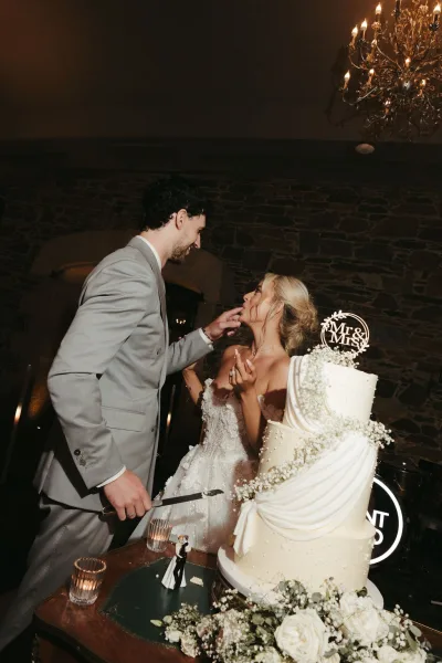 Cake cutting shifts into a cake feeding moment as the couple stand by a three-tier white cake with a Mr & Mrs topper before a stone wall.