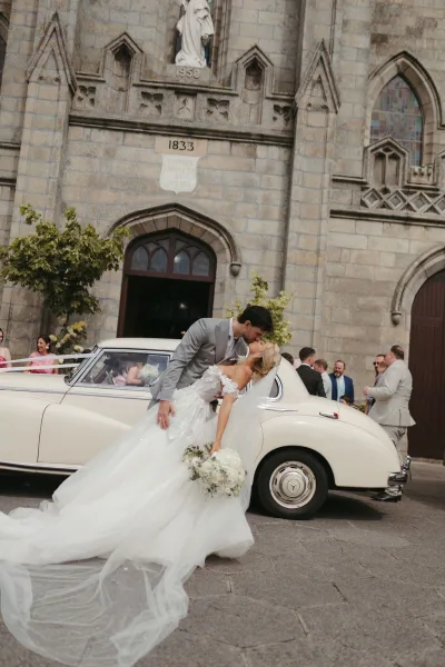 Wedding kiss portrait capturing a dip kiss moment as newlyweds embrace beside a classic car outside a stone church with stained glass.