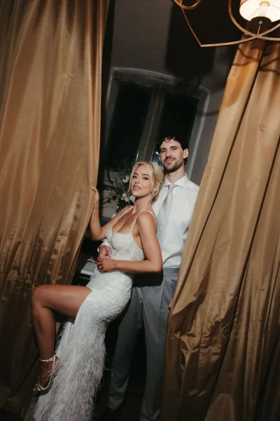 Couple portrait with the bride in a sparkly bridal gown and feather hem, standing with the groom before gold curtains and a chandelier.