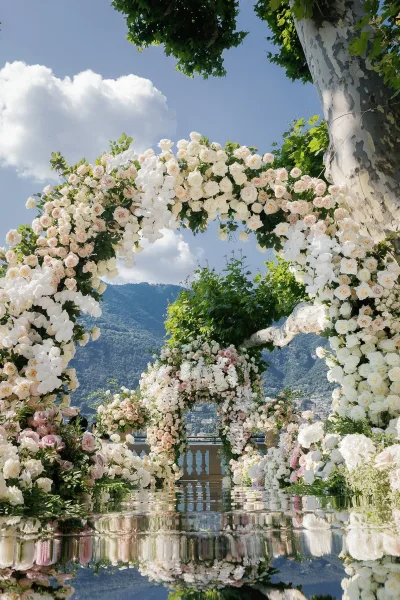 Ceremony arch with a floral wedding arch of roses, white orchids, and greenery, framing a reflective aisle on a terrace with lake and mountain view