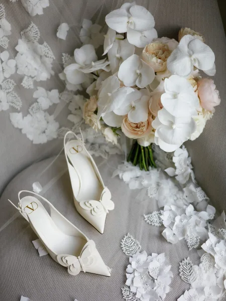 Bridal details flatlay with white slingback wedding heels, lace veil, and orchid-rose bouquet on a gray fabric backdrop