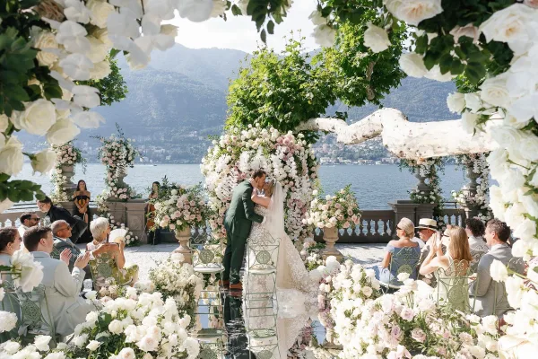 Ceremony kiss during an outdoor wedding ceremony under a round floral arch of white and blush roses on a stone terrace by a mountain lake