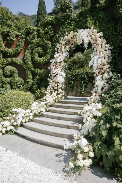 Ceremony entrance decor with a wedding floral arch of white orchids, blush roses, and greenery on stone steps by an ivy wall