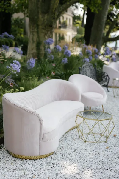 Wedding lounge area with white velvet sofa and chair around a gold geometric coffee table on white gravel, set among garden trees and purple flowers