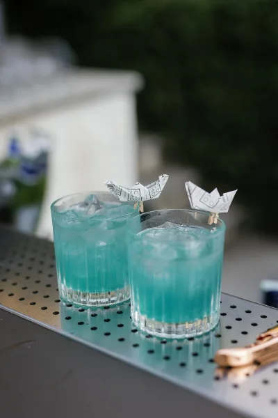Wedding signature cocktails, blue wedding cocktails served on ice in rocks glasses with paper boat toppers on an outdoor bar counter with greenery