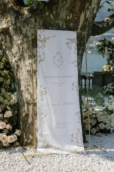 Wedding welcome sign on a gold frame with blush roses and greenery at a lakefront garden entrance beside a tree on white gravel