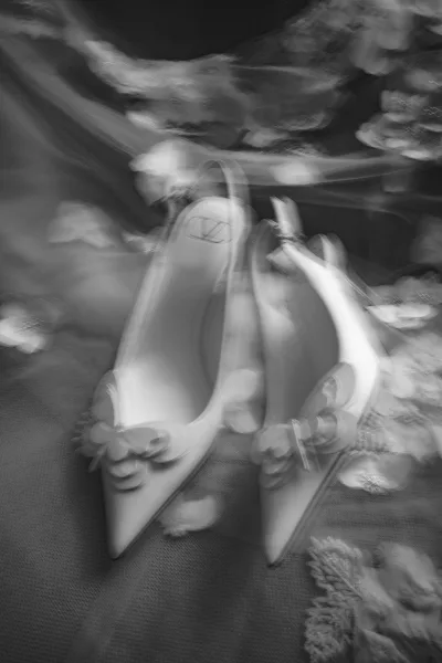 Bridal shoes in a wedding shoe flatlay, white pointed-toe bow heels with designer logo, scattered flower petals on textured fabric