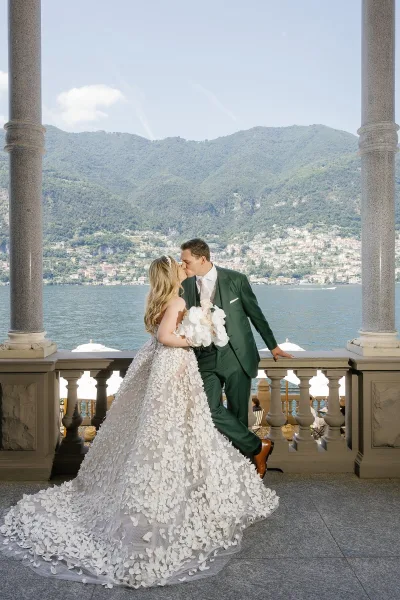 Wedding kiss portrait of bride and groom kiss on a stone balcony, her strapless lace train and white bouquet against lake and mountains.
