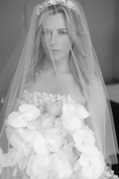 Bridal portrait in black and white of a bride with veil over her face, wearing a floral crown and holding an orchid and rose bouquet indoors
