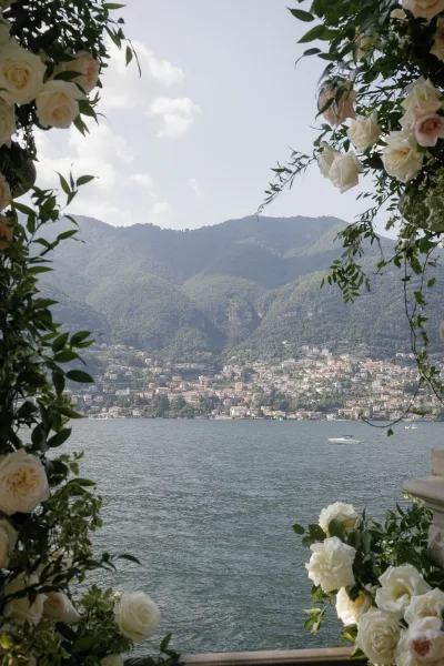 Ceremony arch view with wedding arch flowers, white roses and greenery on a stone terrace overlooking a lake, mountains, and hillside town