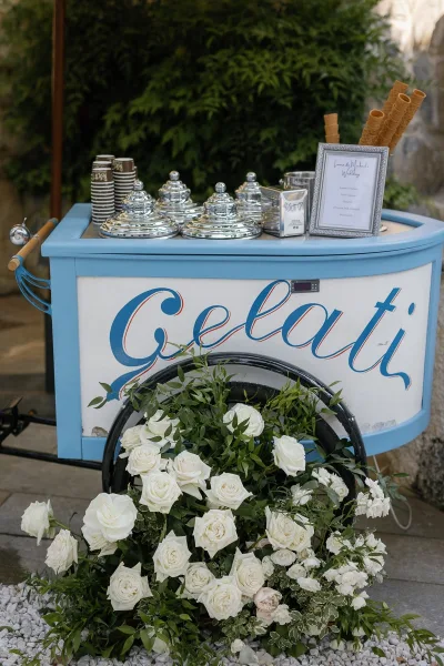Gelato cart at a wedding with framed menu sign, waffle cones and paper cups, plus a floral arrangement beside a wagon wheel on gravel near a stone wall