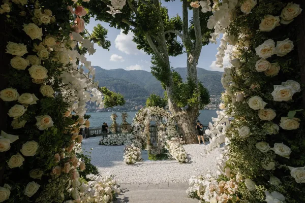 Ceremony setup with a floral ceremony arch, mirrored aisle, and white rose and blush florals on a stone terrace by a mountain lake