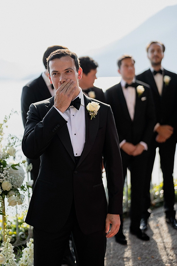 Groom portrait capturing an emotional groom reaction, covering his mouth in a black tuxedo with white rose boutonniere by the ocean aisle florals