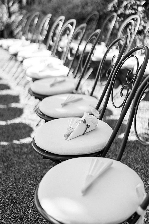 Ceremony seating with outdoor ceremony chairs, wrought iron and white cushions lined along a sunlit gravel path with petal cones