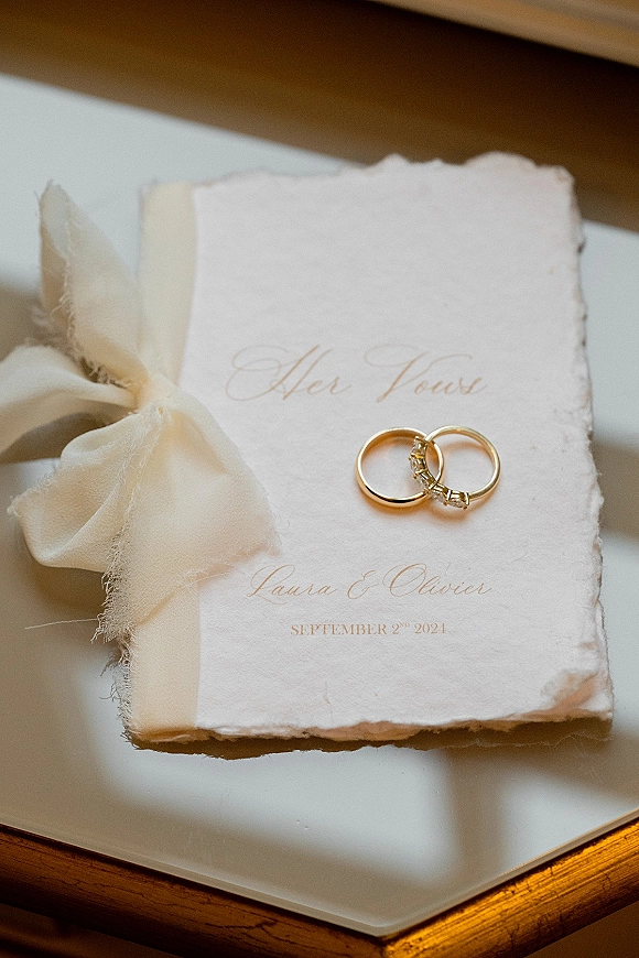 Wedding rings in a wedding ring flatlay, gold bands with a diamond band atop a vow book tied with silk ribbon on a wood table