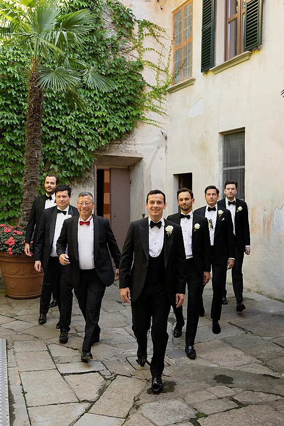 Groomsmen portrait with black tuxedos and bow ties, walking in a stone courtyard beside an ivy-covered wall and stucco building