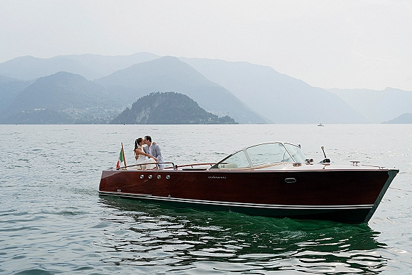 Wedding couple portrait of bride and groom on boat in a classic wooden speedboat with Italian flag, lake and mountains in hazy sky