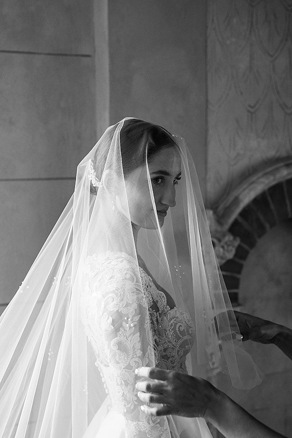 Bridal portrait in black and white of a bride wearing veil, looking over her shoulder in a lace gown by an indoor fireplace mantel