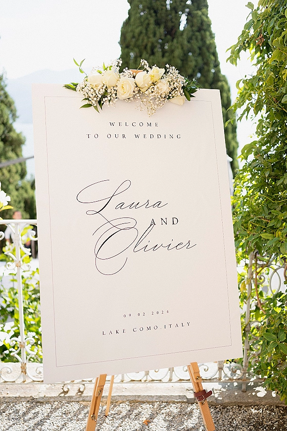 Wedding welcome sign with calligraphy lettering on a wooden easel, topped with white roses and greenery, set by cypress trees and a lake view