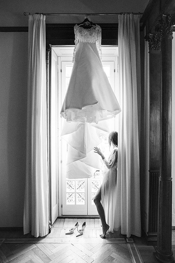 Wedding dress hanging photo of a lace wedding dress on a hanger by a tall window with sheer drapes, above bridal shoes on hardwood floor