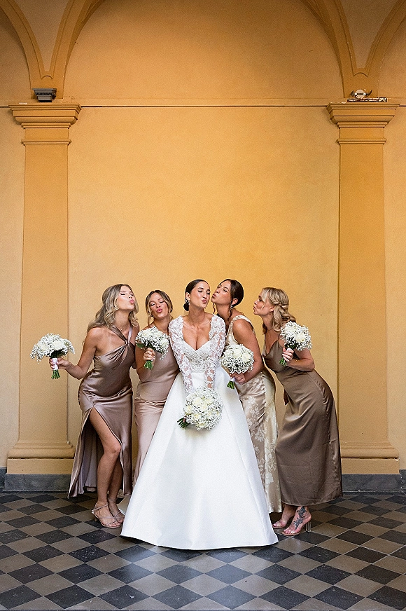 Bride with bridesmaids laughing as they kiss her cheeks, holding white rose bouquets on a checkered tile floor with arched columns