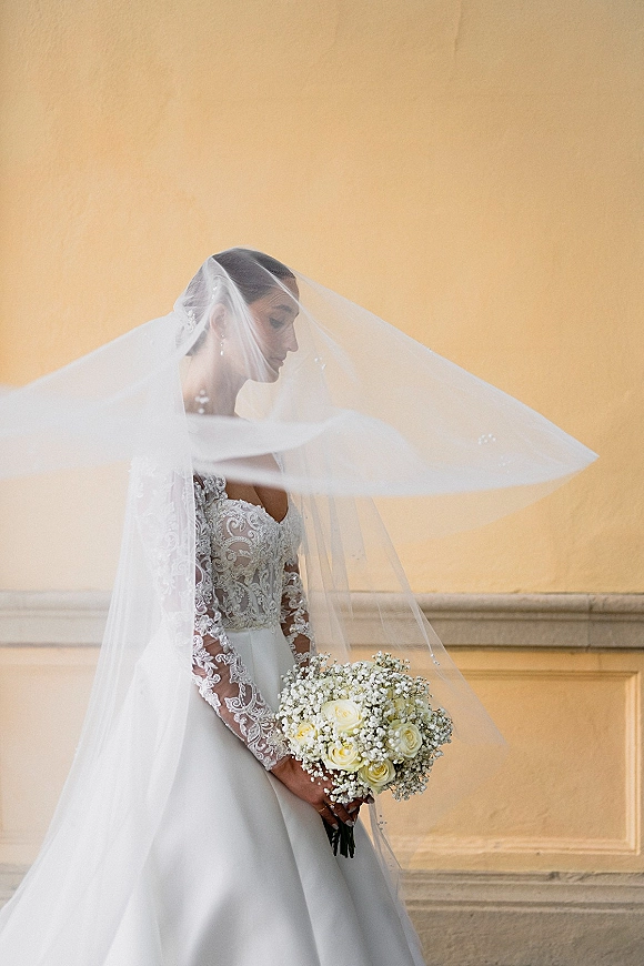 Bridal portrait of a bride with cathedral veil and lace long sleeve wedding dress holding white rose bouquet, near neutral wall molding