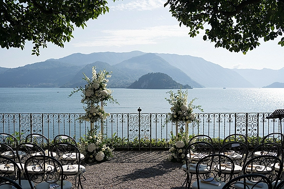 Ceremony setup for an outdoor wedding ceremony with wrought iron chairs and white floral pillars on a terrace overlooking a lake and mountains