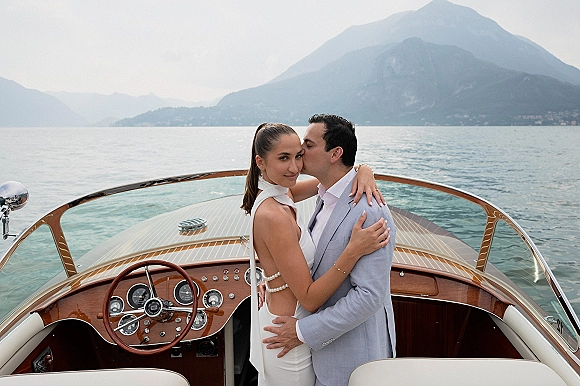 Couple portrait of groom kissing bride in a wedding dress with pearl body jewelry on a speedboat deck, lake and mountains under cloudy sky