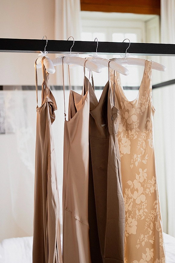 Bridesmaid dresses in neutral tones hang on dress hangers, satin slips and a floral print, lit by a bedroom window near the bed