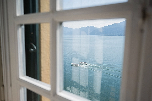 Wedding venue view with a window frame accent overlooking a calm blue lake, boats leaving a wake, and mountains under open sky