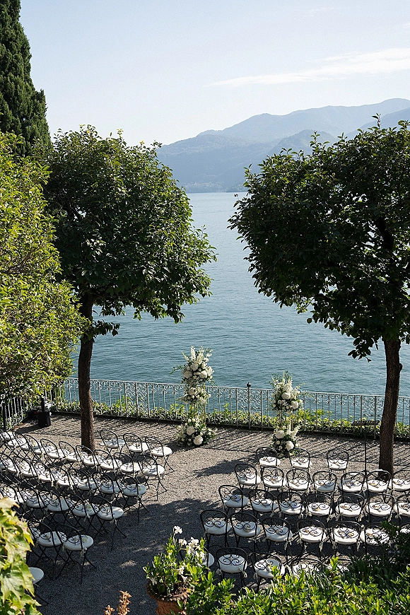 Ceremony setup with curved wrought iron chairs and white floral pillars on a terrace overlooking a lake and mountain backdrop