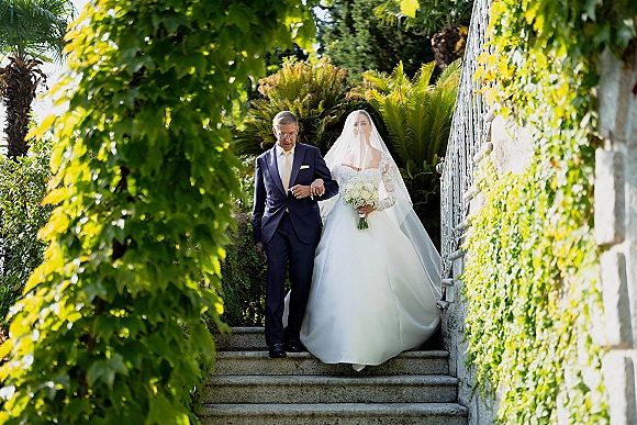 Wedding processional with bride walking down stairs in long sleeve lace gown and veil, holding white rose bouquet beside groom on ivy-lined stone steps