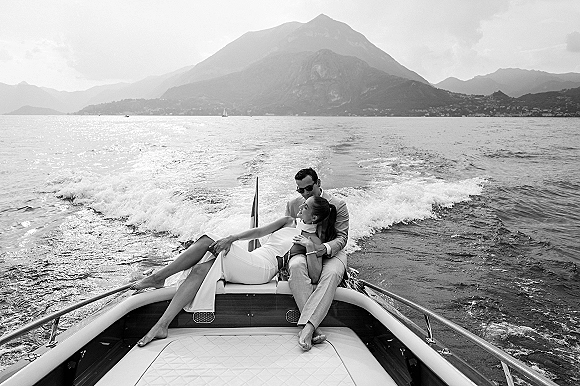 Couple portrait of newlyweds on a boat, bride in strapless dress and sunglasses with groom in suit on a speedboat by a mountain lake shoreline