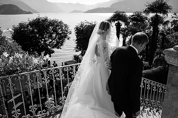 Wedding processional as bride walking down stairs with long cathedral veil and lace back dress, father at her side on a lakeside terrace