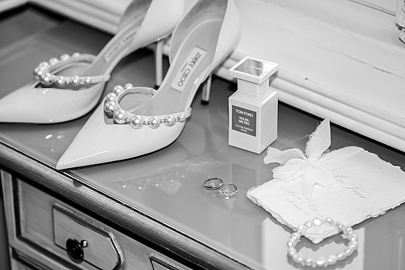Wedding details flatlay with bridal accessories flatlay, pearl strap heels, perfume, rings, pearl bracelet, and ribbon vow book on dresser top