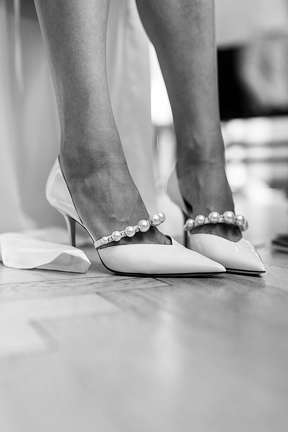 Bridal shoes with pearl wedding heels straps, white pointed-toe pumps set on a wood floor in an indoor room with blurred furniture