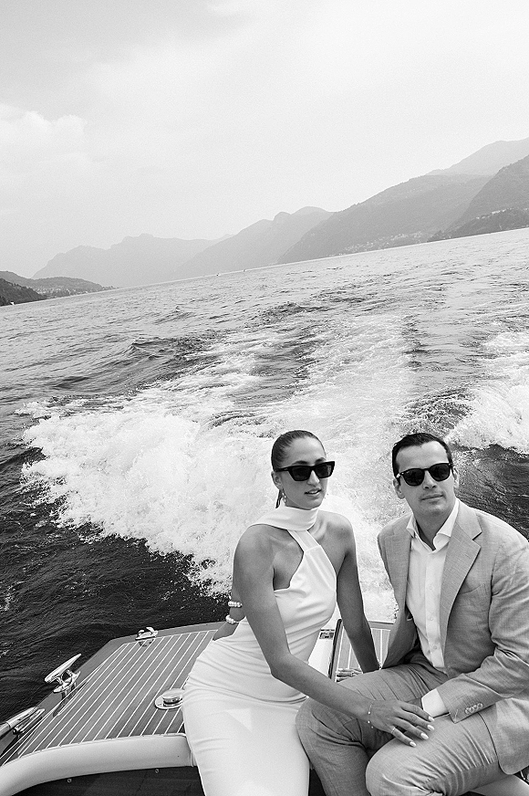 Couple portrait of bride and groom on boat, wearing sunglasses as the boat cuts a wake across a mountain lake under open sky