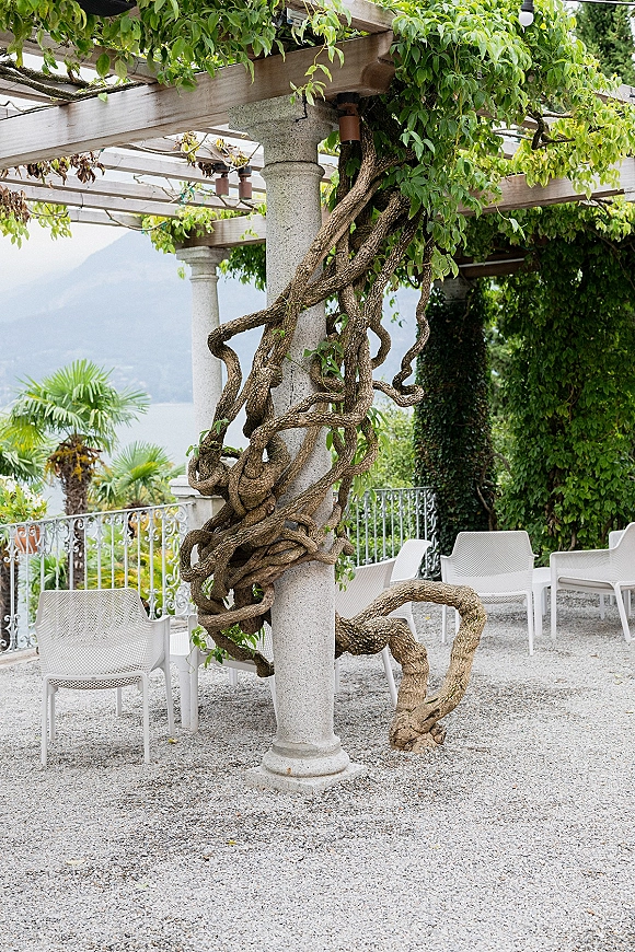 Outdoor wedding venue with a garden pergola wedding setup, vine-covered stone columns and white chairs facing a lakeside mountain view