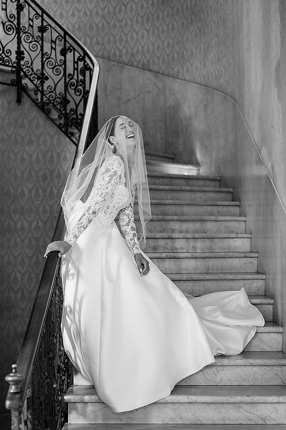 Bridal portrait of a bride on staircase in a long sleeve lace wedding dress and veil, laughing by wrought iron railing and marble steps