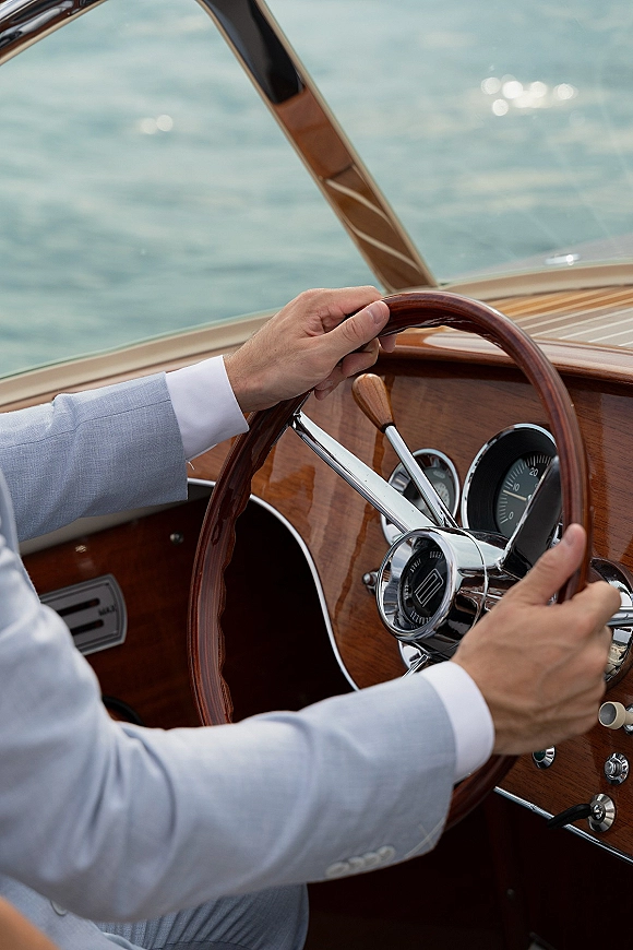 Groom driving boat grips a wooden steering wheel, light blue suit cuffs visible beside chrome gauges on a varnished console, water beyond