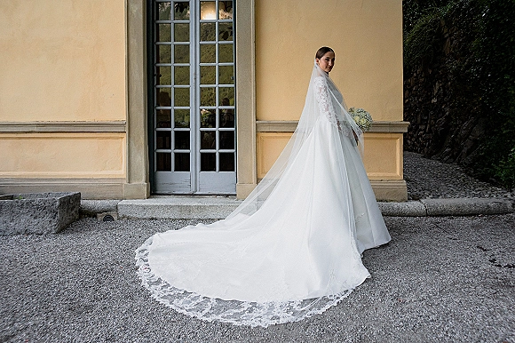 Bridal portrait of a bride in a wedding dress train with lace train and long veil, holding a bouquet by stucco doors on a stone path.