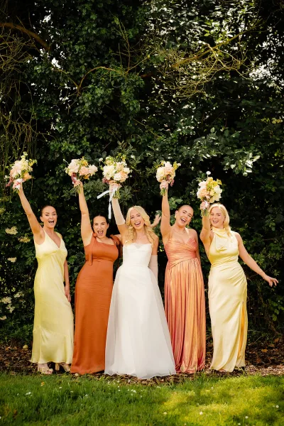 Bridesmaid group photo with bride and bridesmaids holding bouquets, the bride in long gloves on a lush garden lawn under trees