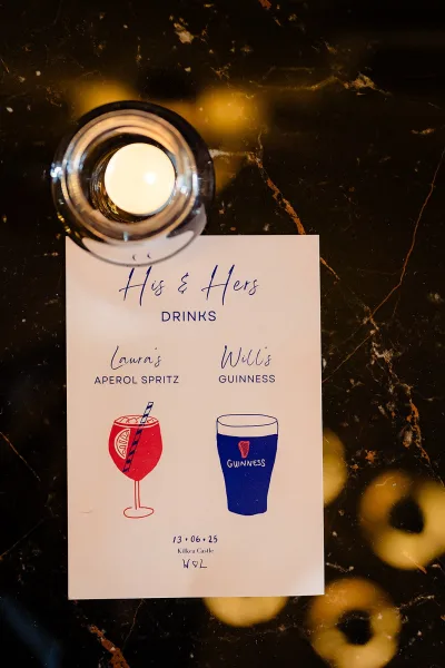 Wedding drink menu and his and hers drinks sign on a dark marble tabletop beside a candle in a glass holder under warm lights