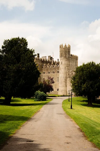 Castle wedding venue exterior with stone turrets and arched windows, flag flying above a paved driveway, manicured lawn under cloudy sky