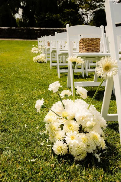 Ceremony aisle decor with white folding chairs and low white floral arrangements along the grass aisle on a green lawn by trees and a stone wall