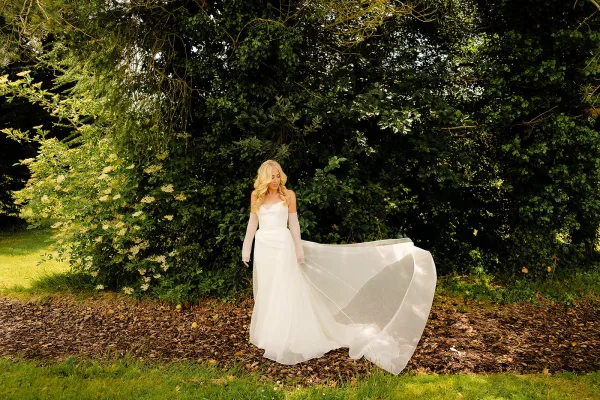 Bridal portrait of a bride in a strapless wedding dress with a long train and sheer gloves, looking down in sunlit garden greenery