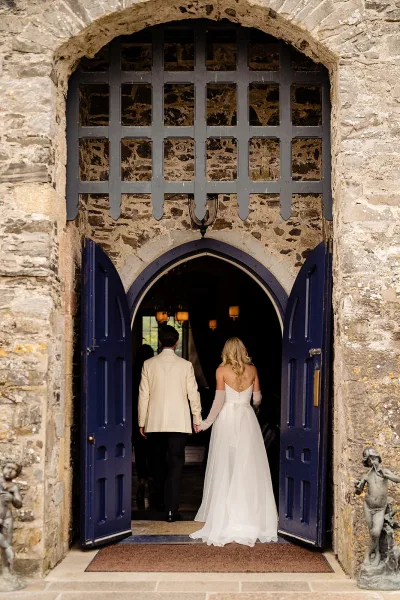 Wedding couple entrance with newlyweds holding hands, walking away into a castle doorway under a stone arch with blue doors and lanterns